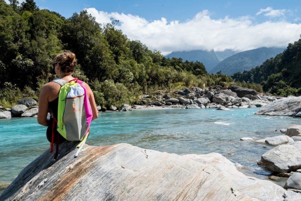 Hiker sitting on large rock near water wearing brightly colored Cotopaxi Luzon daypack